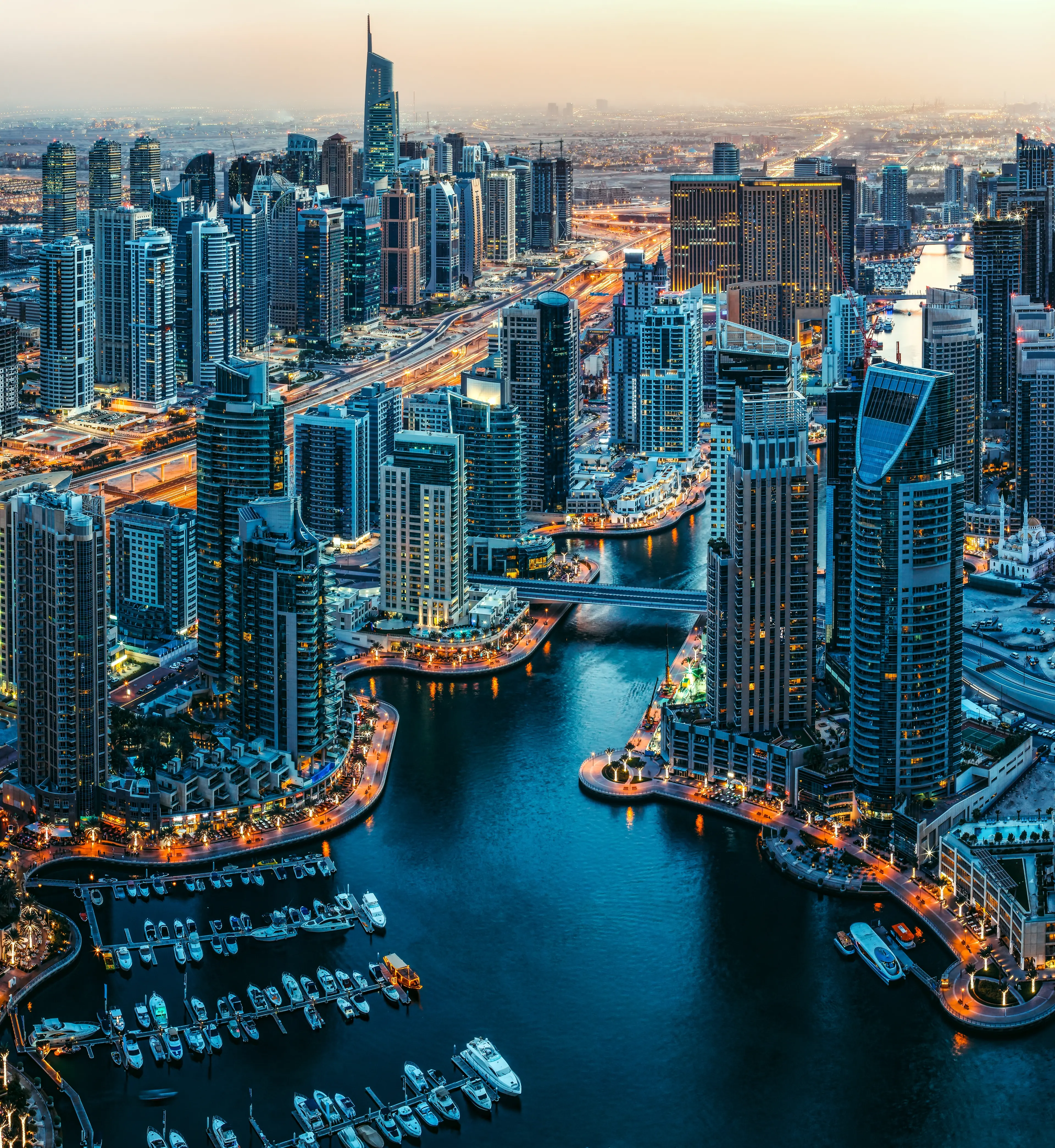 Illuminated skyscrapers and yachts surround a winding blue canal in Dubai Marina at dusk.